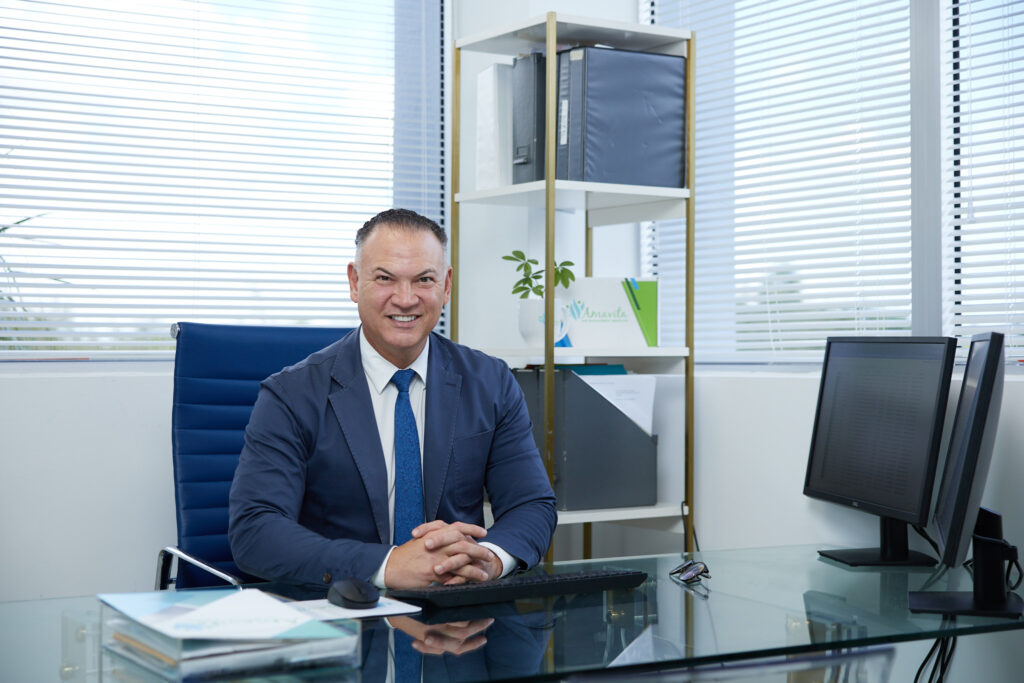 Professional man seated at an office desk, smiling, with company branding visible in the background.